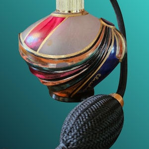 Op de afbeelding: Een sierlijke parfumfles met een goudkleurige dop en een zwarte kwast. De fles heeft een matglazen body met rode, blauwe en gouden accenten. Het ontwerp is elegant en vintage.