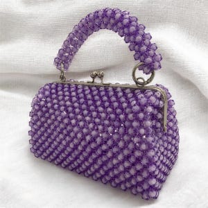 May include: A small, beaded handbag in a vibrant purple hue. The purse features a rounded shape, a silver clasp, and a matching beaded handle. The beads are tightly packed, creating a textured surface. The bag is approximately 7 inches wide.