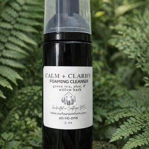 Könnte beinhalten: Eine schwarze Flasche mit Reinigungsschaum und einem transparenten Deckel. Das Etikett lautet "CALM + CLARIFY FOAMING CLEANSER" mit Grüntee, Aloe Vera und Weidenrinde. Die Flasche ist 59 ml und vor grünem Laub platziert.