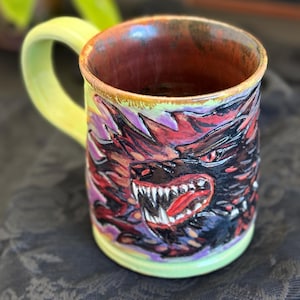 May include: A handmade ceramic mug with a detailed, colorful wolf design. The mug features a light green handle and rim, with a dark red interior. The wolf's face is painted in shades of red, purple, and black, with sharp white teeth.