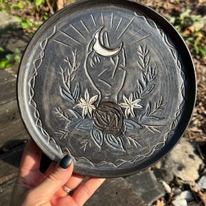 May include: A round, dark gray ceramic plate featuring an embossed design of a hand holding a crescent moon, surrounded by floral elements and leaves. The plate has a decorative wavy border pattern etched along the outer rim of the circular surface.