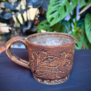 May include: A handmade brown ceramic mug with a textured surface and a decorative leaf pattern. The mug has a curved handle and sits on a dark blue surface. The mug is designed for hot beverages.