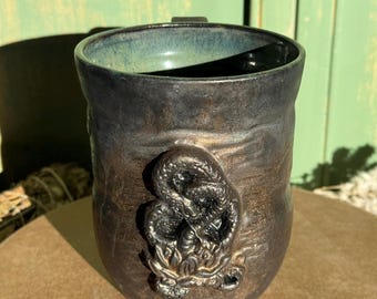 May include: A handmade ceramic mug with a dark, textured exterior and a teal interior. A detailed, raised dragon design adorns the front. The mug has a handle and is suitable for hot or cold beverages.
