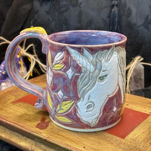 May include: A ceramic mug with a glossy purple and pink glaze featuring a raised white unicorn head relief on the front. The design includes embossed star shapes and yellow leaf accents. The handle is curved and matches the purple and pink glaze.