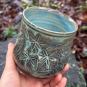 May include: A ceramic mug featuring a dark brown base with carved floral patterns and a light blue glazed interior. The exterior surface displays intricate botanical engravings of star-shaped flowers and leaves, with a smooth, rounded rim and body.