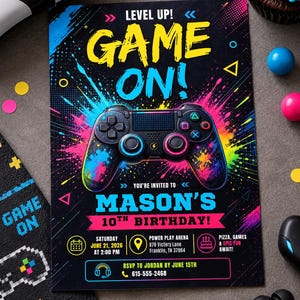 Puede incluir: Una tarjeta de invitación de cumpleaños rectangular con fondo negro, salpicaduras de pintura neón, un gráfico central de mando de juego y el texto: LEVEL UP! GAME ON! MASON'S 10TH BIRTHDAY! SATURDAY JUNE 21, 2026 AT 2:00 PM, POWER PLAY ARENA, 876 VICTORY LANE, FRANKLIN, TN 37064.