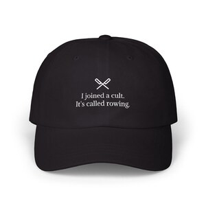 May include: Black baseball cap with a curved brim. The cap features white text that reads "I joined a cult. It's called rowing." Above the text is a white graphic of crossed oars.
