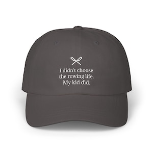 May include: A charcoal gray baseball cap with a curved brim. The cap features white text that reads "I didn't choose the rowing life. My kid did." Above the text is a white graphic of crossed oars.