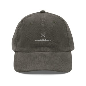 May include: A corduroy baseball cap in a muted olive green color. The cap features a small embroidered design of crossed oars above the word "extraOARdinary" in white lettering. The cap has a curved brim and a rounded crown.