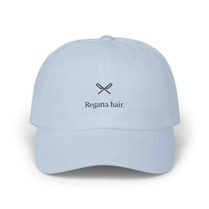May include: A light blue baseball cap with a curved brim. The cap features the text "Regatta hair." in a black, sans-serif font, along with a small graphic of crossed oars above the text. The cap is made of a soft material.