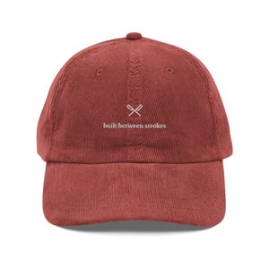 May include: A rust-coloured corduroy baseball cap with a curved peak. The cap has the text "built between strokes" in white, and a small graphic of crossed oars. The hat is a classic six-panel design.