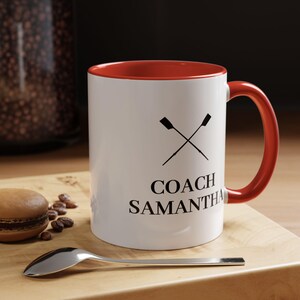 Peut inclure: Mug en céramique blanche avec intérieur et anse rouges. Le mug présente des rames croisées et le texte "COACH SAMANTHA" en noir. Une cuillère et un macaron sont sur une surface en bois.