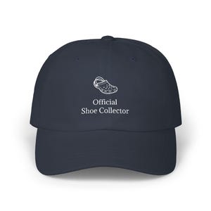 May include: Navy blue baseball cap with a curved brim. The cap features a white graphic of a shoe and the text "Official Shoe Collector" in white. The hat is made of a soft material and has a structured crown.