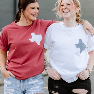 May include: Two people wearing t-shirts with a Texas outline graphic. One shirt is a maroon color, the other is white. The person on the left is wearing light blue denim shorts, and the person on the right is wearing black ripped jeans.