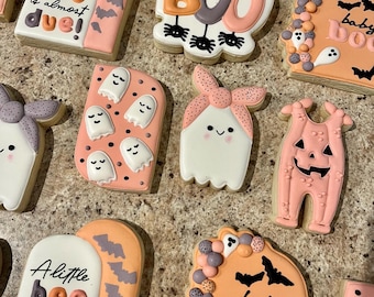 May include: A set of Halloween-themed sugar cookies decorated with pink, white, and black icing. The cookies feature ghosts, bats, and the words "A little boo is almost due!" and "baby boo".