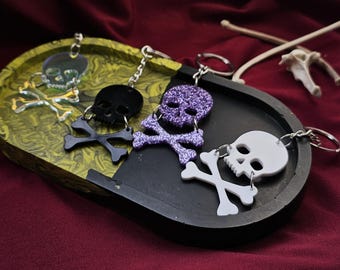 May include: Three skull and crossbones keychains. One is black, one is purple glitter, and one is white. They are all hanging from a black tray.