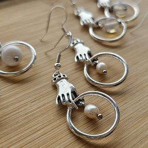 Puede incluir: Pendientes plateados con un diseño de mano que sostiene un anillo con una perla. Los pendientes tienen un cierre de gancho. Los pendientes se exhiben sobre una superficie de madera. Una joya única.