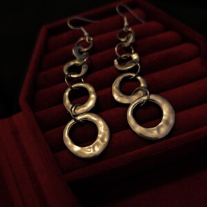 Puede incluir: Un par de pendientes de aro colgantes de plata con múltiples anillos texturizados. Los pendientes se muestran en una caja de joyería de terciopelo rojo.