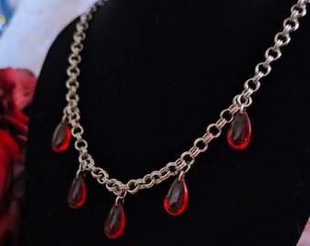May include: A silver-toned chain necklace with five red teardrop-shaped pendants. The necklace is displayed on a black mannequin. The pendants are a vibrant red color and appear to be made of glass or a similar material.