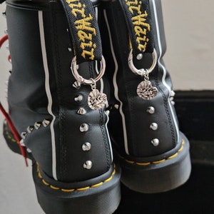 May include: Black leather boots with silver studs and white stripes. The boots have yellow stitching and a black pull tab with yellow text. Silver heart charms hang from the pull tabs. A silver chain bracelet is visible.