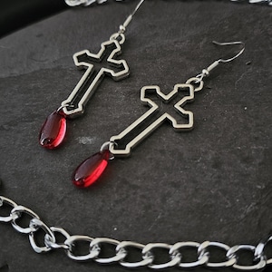 May include: A pair of silver cross earrings with red teardrop-shaped accents. The earrings are hanging from a silver chain.