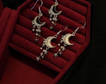 May include: A pair of silver crescent moon earrings with dangling stars. The earrings are displayed in a red velvet jewelry box.