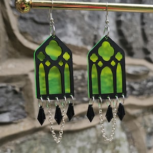 Può includere: Un paio di orecchini in acrilico nero e verde. Gli orecchini hanno la forma di finestre gotiche con un interno verde e un contorno nero. Catene d'argento e ciondoli neri a forma di diamante pendono dal basso.