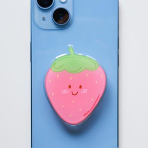 Op de afbeelding: Een roze aardbei telefoonhouder met een lachend gezicht en groene bladeren is bevestigd aan een lichtblauwe smartphone. De telefoon heeft een camera met dubbele lens.