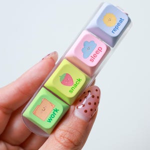 Puede incluir: Una funda de plástico transparente que contiene cuatro teclas de teclado de colores. Cada tecla tiene un color diferente y una palabra: "work", "snack", "sleep" y "repeat". Las teclas también presentan ilustraciones de una carpeta, una fresa, una nube y un sol.