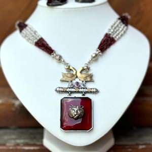 May include: A silver and gold necklace with a red gemstone pendant featuring a lion's head. The pendant is surrounded by a silver frame and is attached to a silver chain with two gold peacocks. The necklace is made of red and white beads.
