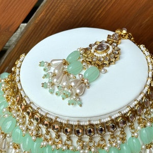 May include: A gold and white necklace with green beads and a matching pendant. The necklace has multiple layers of beads and is decorated with small, clear stones.