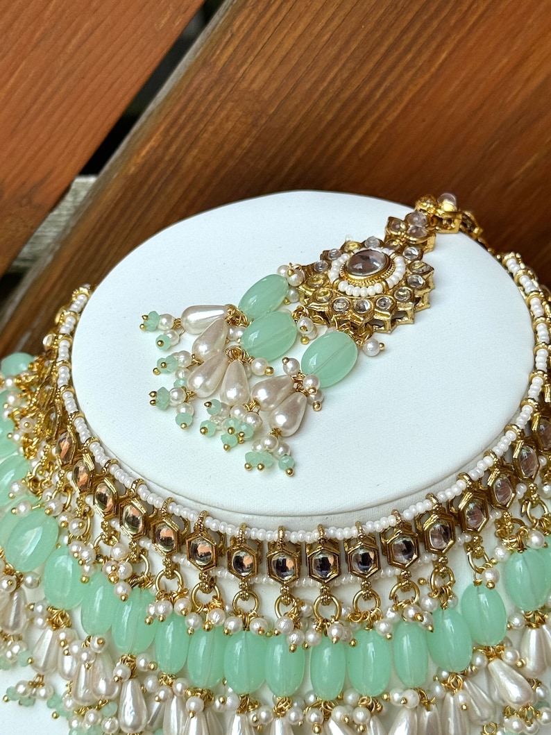 May include: A gold and white necklace with green beads and a matching pendant. The necklace has multiple layers of beads and is decorated with small, clear stones.