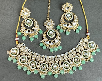 Op de afbeelding: Een gouden ketting, oorbellen en een tikka-set met groene kralen en heldere stenen. De ketting heeft een gelaagd ontwerp met meerdere rijen kralen en stenen.
