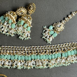 May include: A gold and green Indian bridal jewelry set with a choker necklace, earrings, and a maang tikka. The set features intricate designs with pearls and green stones.