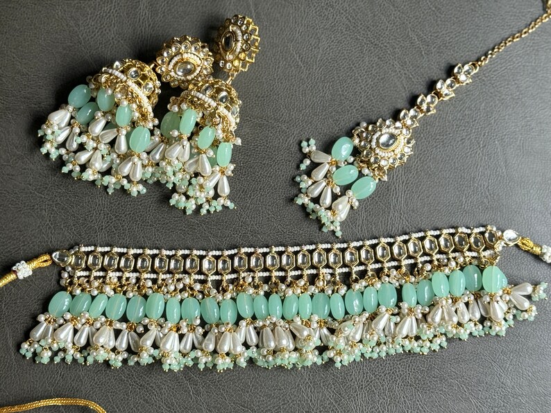 May include: A gold and green Indian bridal jewelry set with a choker necklace, earrings, and a maang tikka. The set features intricate designs with pearls and green stones.