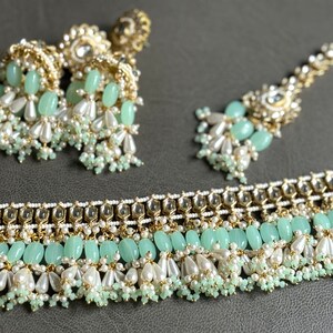 May include: A set of green and gold Indian jewelry featuring a necklace, a pair of earrings, and a tikka. The jewelry is adorned with pearls, beads, and green stones.
