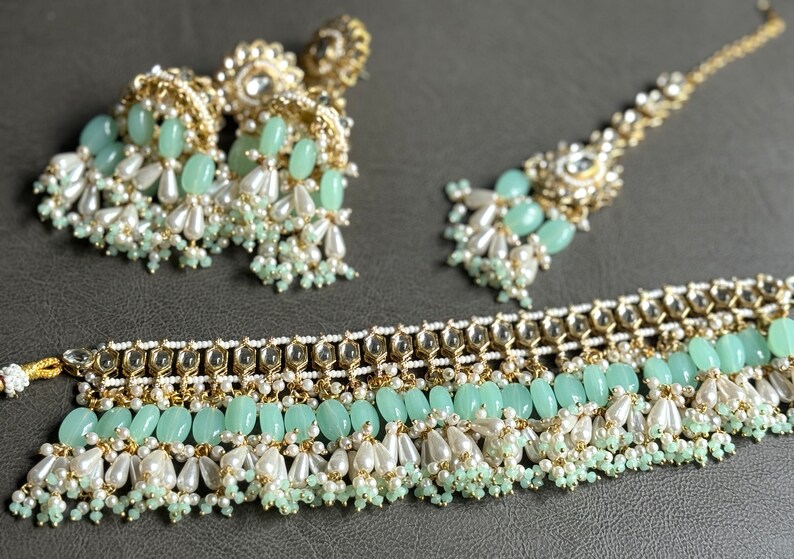 May include: A set of green and gold Indian jewelry featuring a necklace, a pair of earrings, and a tikka. The jewelry is adorned with pearls, beads, and green stones.