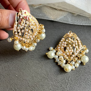 May include: A pair of gold earrings with intricate designs and white pearl accents. The earrings have a teardrop shape and are adorned with small pearls.