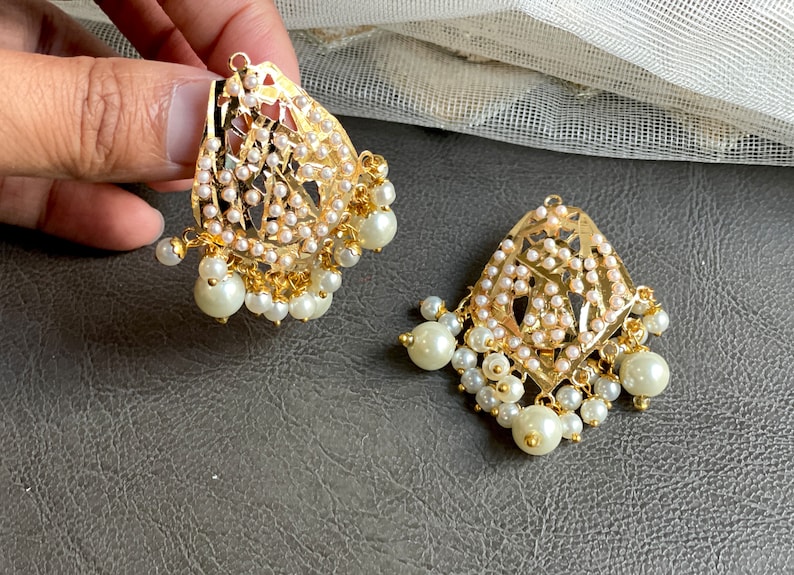 May include: A pair of gold earrings with intricate designs and white pearl accents. The earrings have a teardrop shape and are adorned with small pearls.
