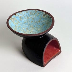 May include: A handcrafted ceramic bowl with a unique design. The bowl features a light blue interior with speckled detailing and a dark brown rim. It sits atop a black, curved base with a red interior. A modern, artistic piece.