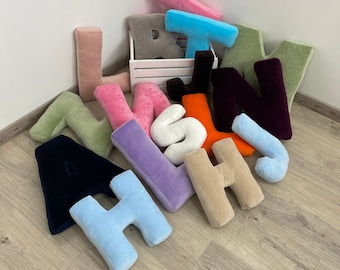 May include: A collection of plush alphabet letters in various colors, including pink, blue, green, purple, and black. The letters are arranged in a pile on a wooden floor.