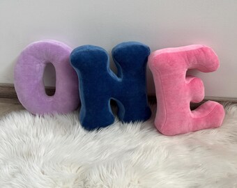 May include: Three plush letters spelling "ONE". The letter "O" is lavender, the "H" is navy blue, and the "E" is pink. The letters are on a white, fluffy rug.