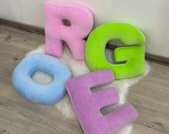 May include: Four plush letters, R, G, O, and E, in pink, green, light blue, and purple, respectively. The letters are arranged on a white fluffy surface.