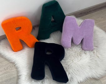 May include: Four plush, oversized letter pillows spelling "RAMR". The letters are in vibrant colors: orange, dark green, lavender, and black. The pillows are arranged on a white, fluffy rug.
