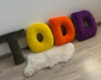 May include: Four plush, colorful letter pillows spelling "TODD" are arranged against a white wall and a wooden floor. The letters are olive green, yellow, orange, and purple. A small, white, fluffy rug is beneath the letters.