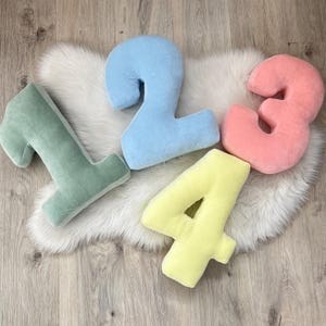 May include: Four plush number pillows in different colors. The numbers are 1, 2, 3, and 4. The pillows are light green, light blue, light pink, and yellow.