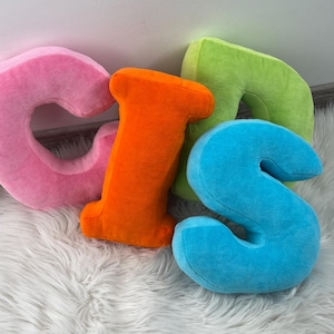 May include: Four plush, colorful letters spelling "G I S N" are arranged on a white, fluffy rug. The letters are pink, orange, green, and blue. The letters are soft and appear to be made of a plush material.