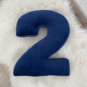 May include: A large, blue, plush number two pillow. The pillow is on a white, fluffy surface.