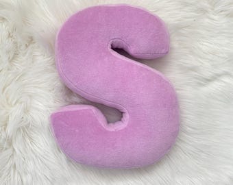 May include: A lavender-colored plush letter "S" pillow. The pillow is made of a soft, velvety material and is placed on a white, fluffy surface. The letter is in a cursive style.