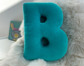May include: A turquoise blue plush letter B pillow with two holes in the shape of the letter B. The pillow is sitting on a white fluffy rug.